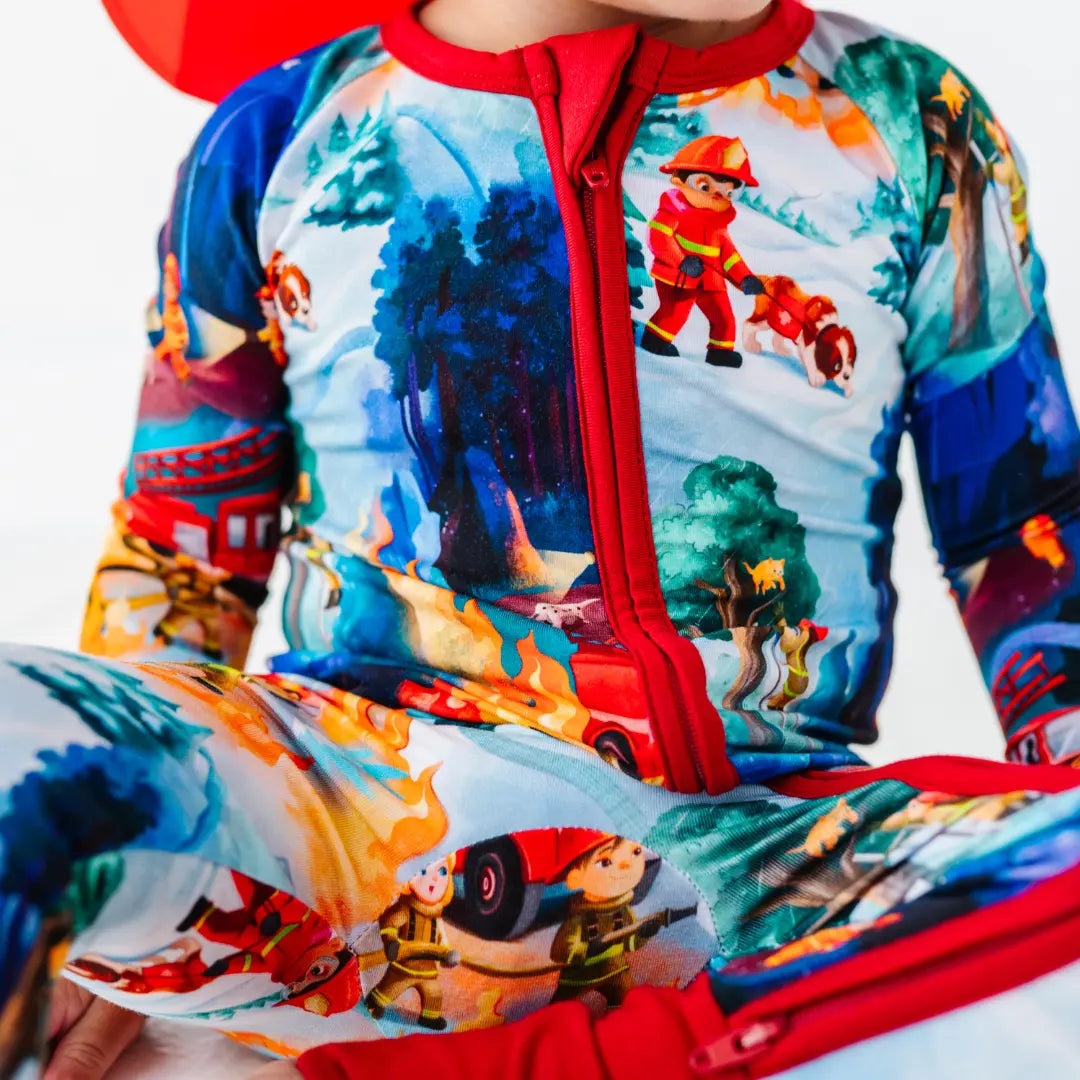 Close-up of a zip-up baby pajama with red trim and a print of firefighters, fire trucks, and flames in a forest scene.